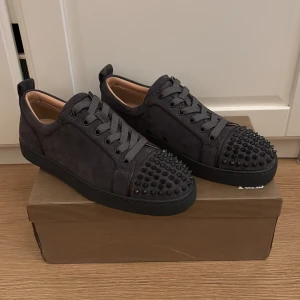 Grå sneakers från Christian Louboutin - Snygga grå sneakers från Christian Louboutin med svarta spikar på tån och klassiska röda sulor. Skorna har snörning och är tillverkade i mocka för en lyxig känsla. Perfekta för att ge din outfit bra detalj. De är helt oanvända och kan gå ner i pris vid snabb affär