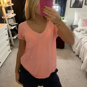 Rosa t-shirt från Gina Tricot - Säljer en söt rosa t-shirt från Gina Tricot i storlek M. T-shirten har en v-ringad hals och en liten bröstficka. Perfekt för en avslappnad stil.