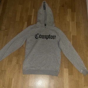 Grå hoodie med 'Compton' tryck - Säljer en grå hoodie med texten 'Compton' tryckt på framsidan. Tröjan va mitt ex så köp den gärna vill inte se den här hemma
