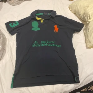 Mörkblå pikétröja från Ralph Lauren - Säljer en mörkblå pikétröja från Ralph Lauren med gröna och orange detaljer. Tröjan har korta ärmar och en klassisk krage. På framsidan finns texten 'Jag Lurar Skatteverket' i grönt.