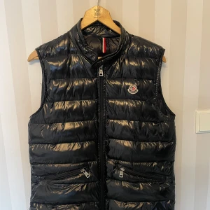 Svart dunväst från Moncler - Snygg svart dunväst från Moncler med glansig yta och dragkedja framtill. Västen har två praktiska fickor med dragkedjor och en broderad logotyp på bröstet. Perfekt för sommar och våren. Dragkedjan lite lös går att fixa!                               Stängs och öppnas utan problem