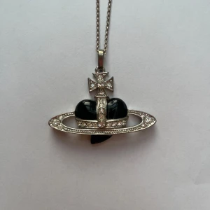 Westwood Saturnus Halsband - Unikt halsband med ett svart hjärta omgivet av en Saturnus ring dekorerad med gnistrande kristaller. Hänget har en distinkt design med en korsdetalj på toppen. Se bild 2 för storleksreferens på smycket.