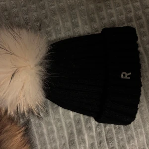  mössa Rock and blue - Snygg svart mössa från Rockandblue med en stor, fluffig pälsboll på toppen. Perfekt för att hålla dig varm och stilren under kalla dagar. Pris kan diskuteras.