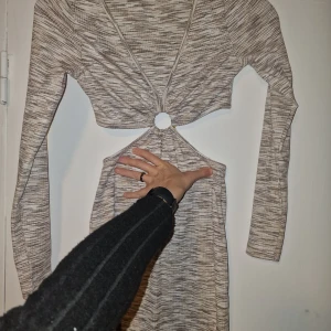 Gråmönstrad klänning med knytning - Trendig gråmönstrad klänning med lång ärm. Klänningen har en unik design med öppen detalj framtill och en rund ring som håller ihop tyget. Perfekt för en stilren look.