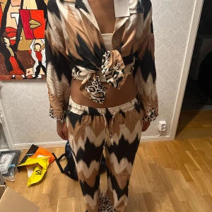 Mönstrad set med knytblus och byxor - Säljer ett snyggt set med en knytblus och matchande byxor. Setet har ett coolt mönster i svart, beige och brunt med leoparddetaljer. Blusen är långärmad och kan knytas framtill för en trendig look. Perfekt för en avslappnad dag eller en utekväll!