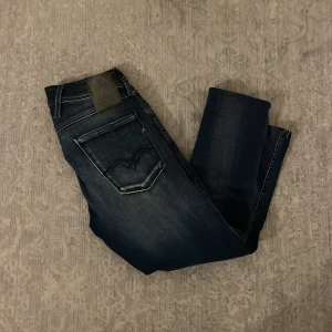 Blå jeans från Replay - Säljer ett par snygga svarta jeans från Replay, modell Anbass. De har en slim fit och är perfekta för både vardag och fest. Jeansen är i mycket bra skick och har en klassisk femficksdesign med dragkedja och knapp. Passar perfekt till en casual eller dressad stil.