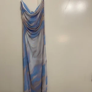 Blå och beige långklänning - Säljer en elegant långklänning i blå och beige med ett vackert draperat mönster. Klänningen är axelbandslös. Materialet är lätt och luftigt, vilket gör den bekväm att bära hela dagen.