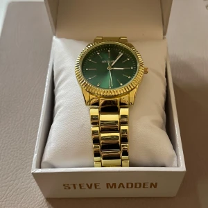 Guldfärgad klocka från Steve Madden - Snygg guldfärgad klocka från Steve Madden med en grön urtavla. Klockan har en stilren design med ett glänsande armband och en räfflad kant runt urtavlan. Perfekt för både vardag och fest!😍