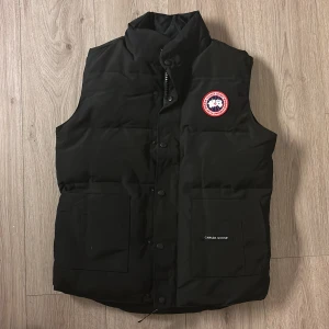 Svart dunväst från Canada Goose - Säljer en svart dunväst från Canada Goose i mycket bra skick. Västen har en hög krage och stängs med både dragkedja och knappar. Den har två stora fickor framtill och det klassiska Canada Goose-märket på bröstet. Perfekt för kyliga höst- och vinterdagar. Storlek xs men den passar som s också.