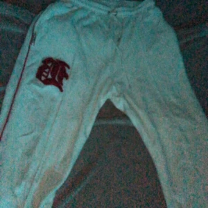 Vita mjukisbyxor med röd detalj - Säljer ett par vita mjukisbyxor med en cool röd broderad detalj på sidan. De har en bekväm passform med snörning i midjan och dragkedja på sidan. Perfekta för en avslappnad dag hemma eller en snabb tur till affären. 🏠 använda en gång dom var för stora