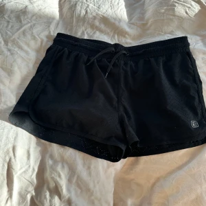 Svarta träningsshorts från Energetics - Säljer ett par svarta träningsshorts från Energetics i mycket bra skick. De är tillverkade i polyester och elastan för en skön och flexibel passform. Perfekta för träning eller löpning med en elastisk midja och dragsko för justerbar passform.