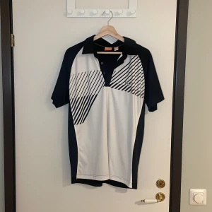 Svart och vit pikétröja från Puma - Säljer en snygg svart och vit pikétröja från Puma i mycket bra skick. Den har ett coolt grafiskt mönster på framsidan och en klassisk krage med knappar. Perfekt för sportiga aktiviteter eller en avslappnad stil. Passar både vår och sommar! Pris går alltid att förhandlas!