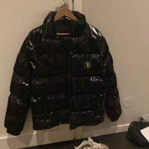 Svart glansig pufferjacka från Trapstar - Säljer en svart glansig pufferjacka från Trapstar. Jackan har en hög krage och en stor huva för extra värme. Den är perfekt för kalla dagar och har en snygg logga på bröstet. Passar både till vardags och festliga tillfällen. Dragkedja framtill och elastiska muddar vid ärmsluten. Säljer eftersom den inte är min still❤️