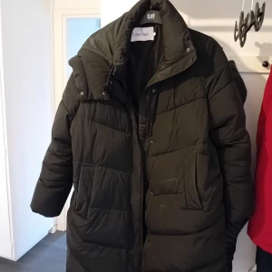 Svart dunjacka från Calvin Klein - Säljer en svart dunjacka från Calvin Klein i mycket bra skick. Den är perfekt för kalla vinterdagar med sin tjocka vaddering och långa ärmar. Jackan har en dragkedja framtill och en hög krage för extra värme. Perfekt för både vardag och festliga tillfällen!
