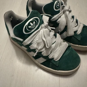 Gröna Adidas Campus sneakers - Säljer ett par gröna Adidas Campus sneakers i bra skick. De har en klassisk design med vita ränder och snörning. Perfekta för vardagsbruk och ger en retro känsla. Skorna är bekväma och stilrena, idealiska för både vår och höst.