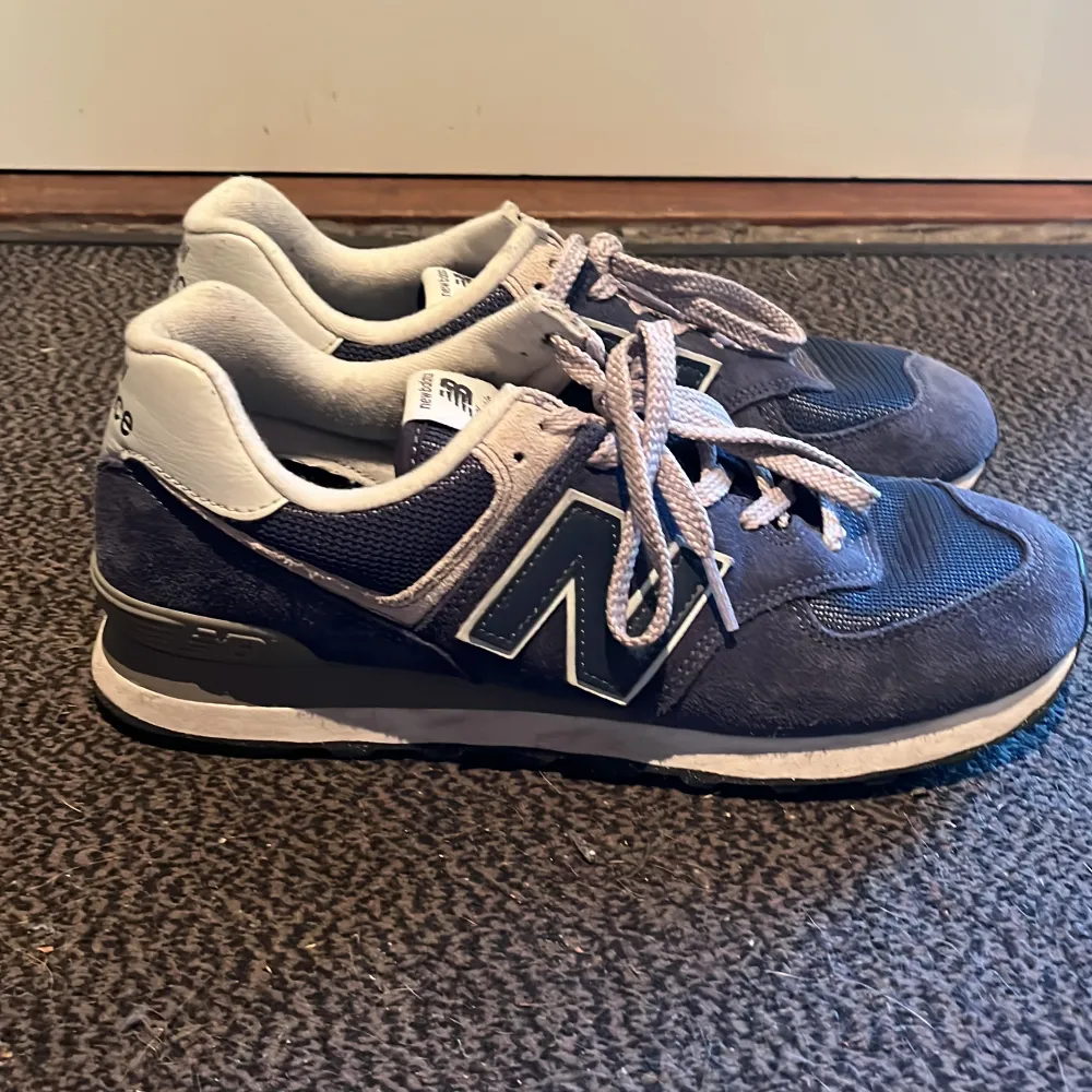 Säljer ett par klassiska New Balance 574 sneakers i blått och vitt. Skorna har en snygg design med snörning och är i bra skick. Perfekta för både vardag och träning. De har en bekväm passform och är gjorda i en kombination av mocka och syntetmaterial.. Skor.