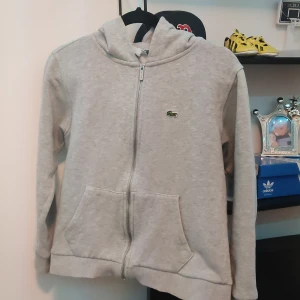 Grå hoodie från Lacoste - Säljer en stilren grå hoodie från Lacoste i mycket bra skick. Den har en dragkedja framtill och den klassiska krokodilloggan på bröstet. Perfekt för både vardag och chill. 🐊