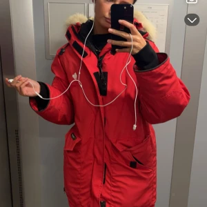 Canada goose jacka - Säljer en snygg röd parkajacka med pälsdetaljer på huvan. Jackan har en dragkedja och knappar framtill, samt praktiska fickor. Perfekt för kalla vinterdagar! 🧥❄️