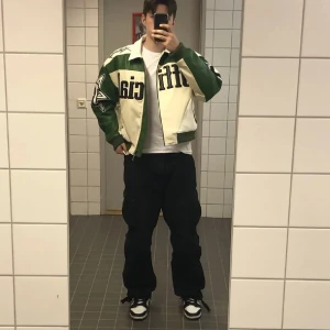 Grön och vit racingjacka från Man Official - Säljer en grön och vit racingjacka från Man Official. Den har en cool design med stora tryck som 'Official' och '73' på framsidan. Jackan har dragkedja och långa ärmar, perfekt för höst, vår och vinter. Den är i bra skick och passar perfekt för en streetwear-look.