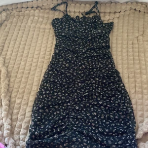 Blommig klänning från Divided - Söt blommig klänning från Divided i svart med små blommor. Den har smala axelband och en tight passform som framhäver figuren. Perfekt för sommarens alla fester eller en mysig dag i parken. Klänningen är i mycket bra skick och redo för nya äventyr!