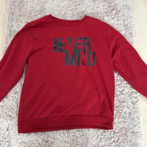 Röd sweatshirt med tryck - Säljer en cool röd sweatshirt med texten 'NEVER MIND' i svart på bröstet. Tröjan har en avslappnad passform och är perfekt för både vardag och chill. Den är i mycket bra skick och redo för en ny ägare. Perfekt för höst och vår!