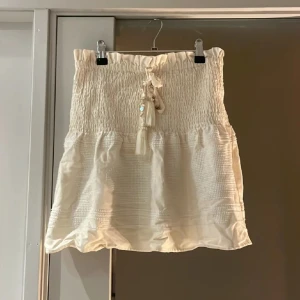 Vit kjol från Zara - Säljer en söt vit kjol från Zara med smockdetaljer och knytband i midjan. Kjolen har en kort längd och är perfekt för varma dagar. Den är tillverkad i ett lätt och luftigt material.