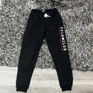 Svarta mjukisbyxor från Vinson Polo Club - Snygga svarta mjukisbyxor från Vinson Polo Club med tryck på benet. Byxorna har en bekväm passform med resår och snörning i midjan samt muddar vid bensluten. Perfekta för en avslappnad stil.