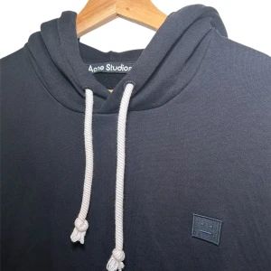 Svart Acne Studios Hoodie - Snygg svart hoodie från Acne Studios med vit dragsko och logotyp på bröstet. Perfekt för en stilren och avslappnad look. Storleken är S men den skulle även kunna passa en M beroende på hur man gillar passformen. Skicket är mycket bra, sparsamt använd