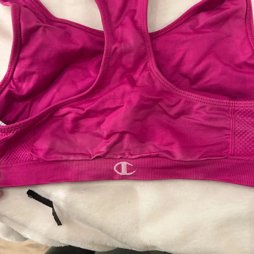 Säljer en rosa sport-bh från Champion i syntetmaterial. Den har en racerback-design och bred resår under bysten för extra stöd. Perfekt för träning och aktivitet.. Yläosat.