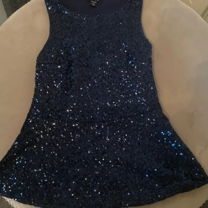 Mörkblå glittrig klänning - Snygg mörkblå klänning täckt med glittriga paljetter. Den är ärmlös och perfekt för en festlig kväll. Klänningen har en elegant passform som framhäver figuren.