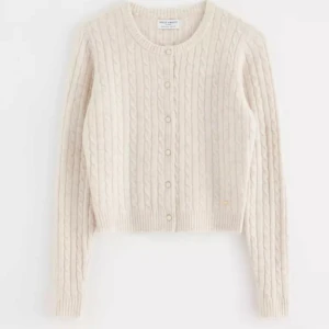 Beige stickad kofta från Holly & White - Säljer en stilren beige stickad kofta från Holly & White. Koftan har ett klassiskt flätmönster och knappar framtill. Perfekt för kyliga dagar med sin mysiga och varma design.