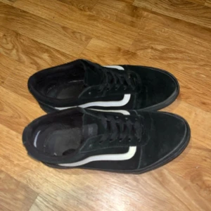 Svarta sneakers från Vans - Säljer ett par klassiska svarta Vans sneakers med vit sidosöm. Skorna har snörning och är tillverkade i mocka. Perfekta för en avslappnad stil.