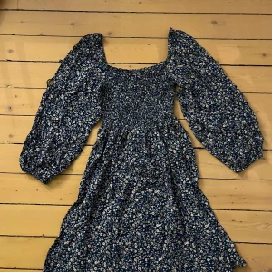 Blommig blå klänning - Gullig blå klänning med små vita blommor. Ärmarna går att ha på axlarna eller ha som off shoulder. Den har en smockad överdel som ger en fin passform. Köpt på en marknad i Spanien. Köpt för 300kr, aldrig använd. Pris kan diskuteras!❤️