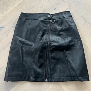 Skinnkjol - Snygg svart kjol i skinnimitation från ONLY med dragkedja framtill och knappdetalj i midjan. Endast använd 1 gång så i mycket bra skick.