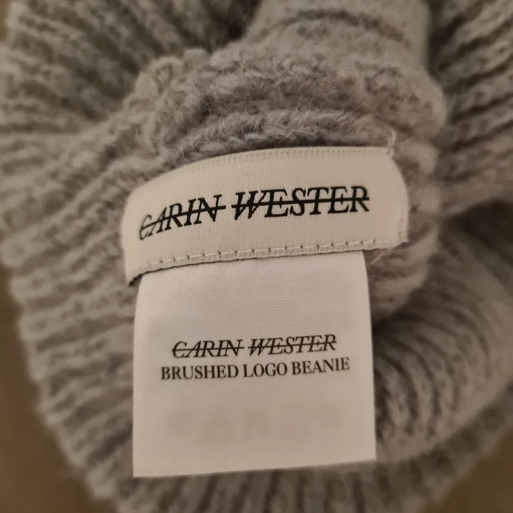 Snygg grå stickad mössa från Carin Wester med ribbad design och ett diskret CW-märke framtill. Perfekt för kyliga dagar och ger en stilren touch till din outfit.. Asusteet.