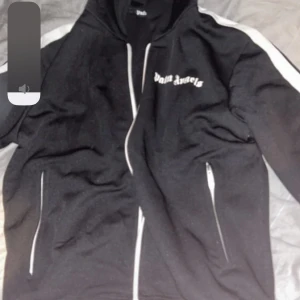 Svart hoodie från Palm Angels - Snygg svart hoodie från Palm Angels med vit text på bröstet. Den har en dragkedja framtill och vita detaljer längs ärmarna. Perfekt för en avslappnad stil.