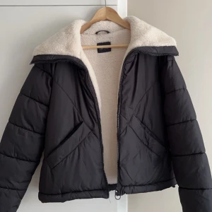 Svart pufferjacka med teddyfoder - Snygg svart pufferjacka med mysigt vitt teddyfoder. Jackan har en dragkedja framtill och en hög krage för extra värme. Perfekt för kyliga dagar när du vill hålla stilen. (EGNA BILDER PRIVAT)