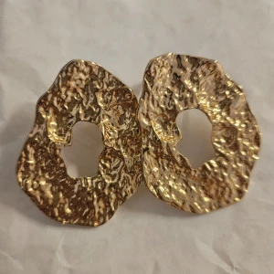 Guldiga abstrakta örhängen - Säljer ett par unika guldiga örhängen med en abstrakt, ojämn form och en glittrig yta. De har en öppen design i mitten och är perfekta för att ge en stilfull touch till din outfit.