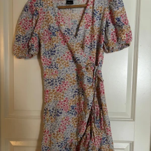 Blommig omlottklänning - Blommig omlottklänning från Gina Tricot med kort ärm och volangdetaljer. Klänningen har en v-ringad design och knytning i midjan samt är en kort modell. Lite slitningar på ett av snörena! Perfekt sommarklänning!!☀️🌸