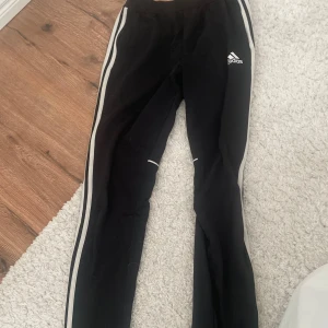Svarta träningsbyxor från Adidas - Snygga svarta träningsbyxor från Adidas med vita ränder längs sidorna. Byxorna har en tight passform och är perfekta för träning. Adidas-loggan är tryckt på ena benet.