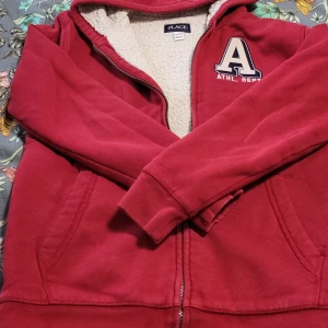 Röd fleecefodrad hoodie från Place - Säljer en röd hoodie från Place med fleecefoder och en stor bokstav 'A' på bröstet. Den har dragkedja och fickor framtill. Hund finns hemma.