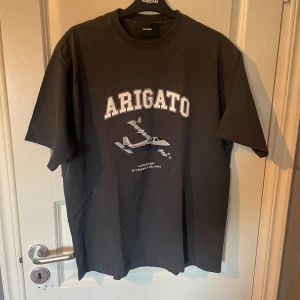 Svart t-shirt från Axel Arigato - Snygg svart t-shirt från Axel Arigato med tryck av ett flygplan och texten 'ARIGATO' på framsidan. Perfekt för en avslappnad stil. Stor i storlek passar M