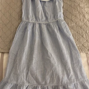 Blå- och vitrandig klänning med volang - Charmig blå- och vitrandig klänning med volangdetalj vid axlarna. Perfekt för sommarens varma dagar. Klänningen har en luftig passform och är tillverkad i ett lätt material.