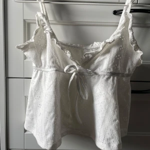 Vit spetslinne med volangdetaljer - Charmigt vitt linne med spets och volangdetaljer. Linnet har smala axelband och en söt knytning framtill. Perfekt för varma sommardagar.