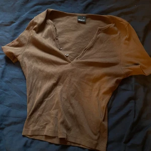 Brun ribbad topp från Gina Tricot - Säljer en brun ribbad topp från Gina Tricot med v-ringning och knappar framtill. Inte använd alls, Toppen har korta ärmar och är perfekt för en avslappnad stil.