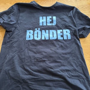 Svart t-shirt med blå text - Cool svart t-shirt med texten 'HEJ BÖNDER' och 'VI É DIF' i blått. På ärmen finns en stiliserad figur med gloria. Perfekt för att visa ditt stöd på matchen eller bara för en avslappnad look.
