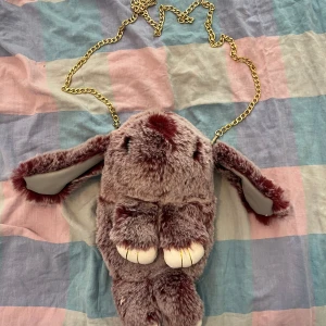 Vinröd pälsväska med kedja - Unik och rolig vinröd pälsväska med guldiga kedjehandtag. Väskan har ett lekfullt djurmotiv med öron och tassar, perfekt för att sticka ut i mängden. Den är mjuk och fluffig, vilket ger en extra touch av stil och humor.