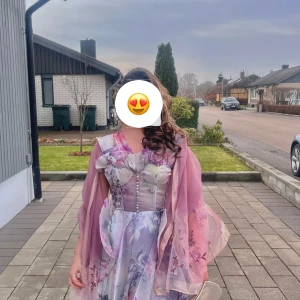 Blommig långklänning med sjal - Elegant långklänning med blommigt mönster i lila och rosa toner. Klänningen har en unik design med en matchande sjal som ger en extra touch av stil. Perfekt för speciella tillfällen.