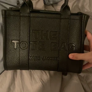Svart tote bag från Marc Jacobs!! - Snygg svart tote bag från Marc Jacobs i skinn. Väskan har två rejäla handtag och en rymlig insida med dragkedja. Perfekt för att bära med sig allt man behöver under dagen. Stilren design med texten ”THE TOTE BAG” på framsidan.