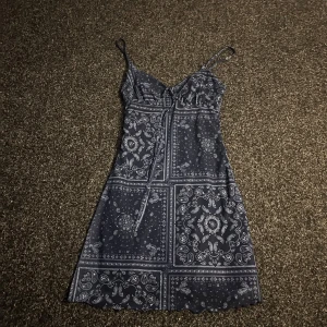 Mönstrad klänning - Snygg blå klänning med ett detaljerat mönster och smala axelband. Klänningen har en knytning framtill och en lätt utställd form. Perfekt för en avslappnad sommarlook.